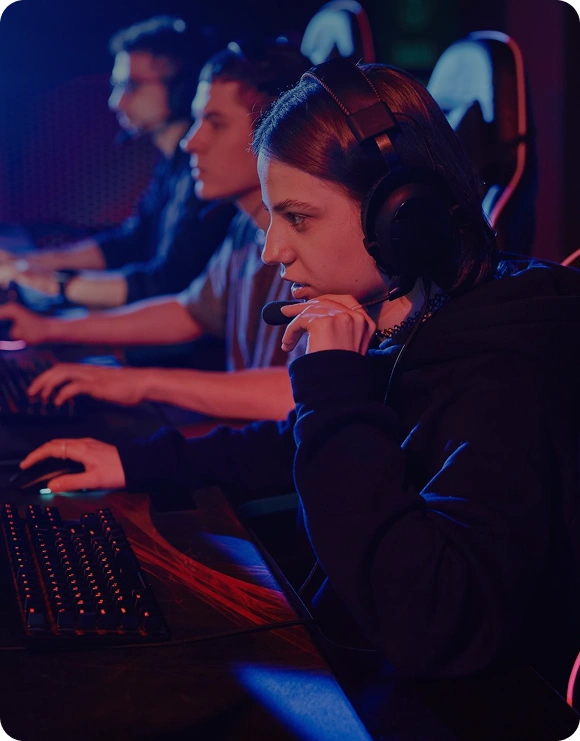 Een speler met headset zit geconcentreerd achter een gamingcomputer tijdens een wedstrijd, terwijl andere spelers op de achtergrond zichtbaar zijn.