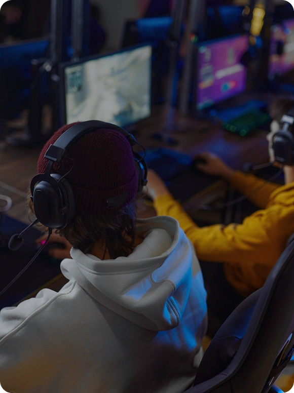Een speler zit met een headset op achter een gamingcomputer in een esports-club en speelt geconcentreerd, terwijl andere spelers op de achtergrond zichtbaar zijn.