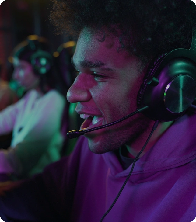 Een esports-speler met headset speelt geconcentreerd achter een gamingcomputer, met andere spelers op de achtergrond in een competitieve setting.