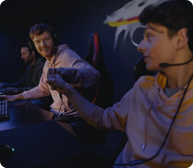 Twee spelers zitten naast elkaar en spelen samen achter gamingcomputers, terwijl ze communiceren en samenwerken tijdens een game.