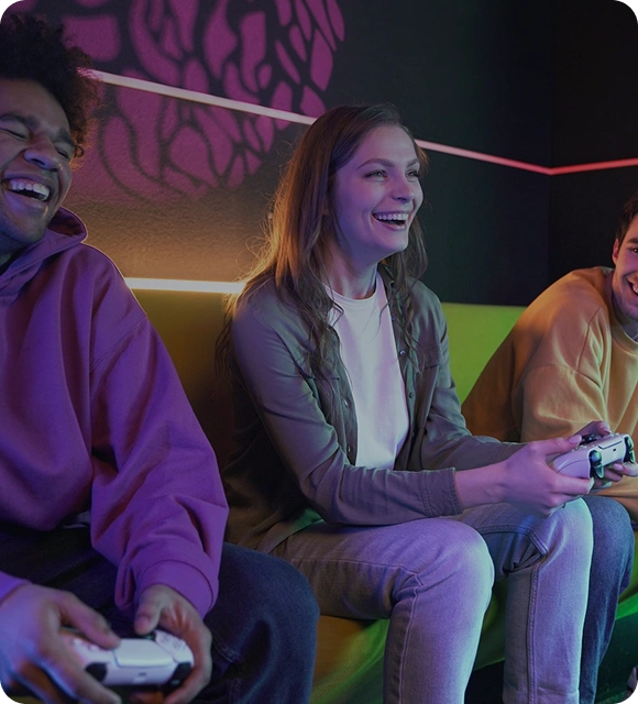 Drie vrienden zitten samen op een bank en spelen een game met controllers, terwijl ze lachen en ontspannen in een gamingomgeving.